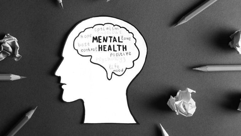Paper cutout of a head with the brain outlined in pen, inside reads "mental health" and positive words, cutout surrounded by pencils and crumpled paper