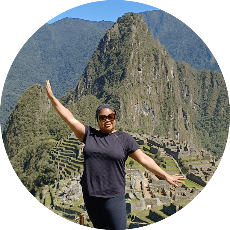Cheyenne wearing sunglasses, a sweatband, and black athletic wear holds her arms wide to show the amazing view behind her of Machu Picchu in Peru.