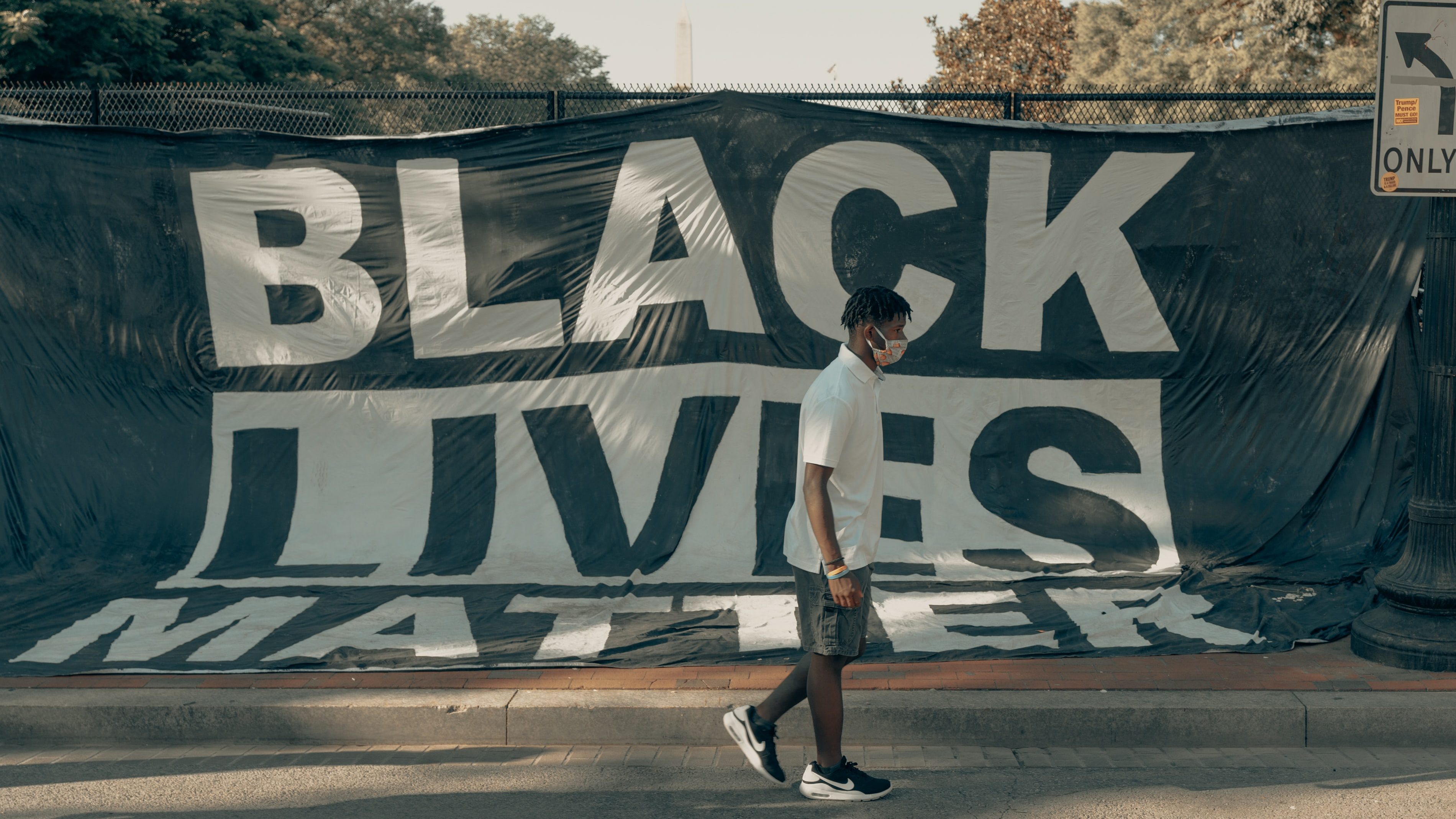Young black man in shorts and sneakers walks in front of a large Black Lives Matter banner on a fence in Washington, DC. Photo by Clay Banks.