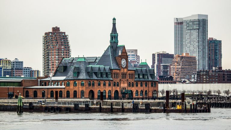 The Central Railroad of New Jersey Terminal in Jersey City from across the water. A few highrises and apartment buildings stand behind it. The Terminal building is red brick with a high arched roof and clocktower. Photo by Gautam Krishnan.