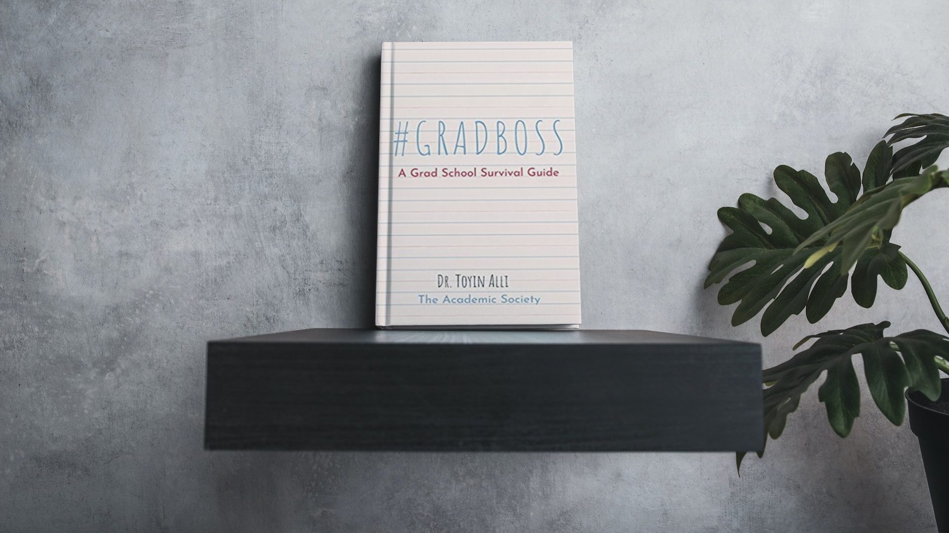 A copy of #GradBoss A Grad School Survival Guide by Dr. Toyin Alli of The Academic Society sits on a black floating shelf next to a potted fern.