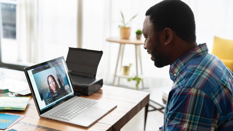 A black man in a plaid button down shirt sits at a desk talking with a woman on video chat on his laptop.
