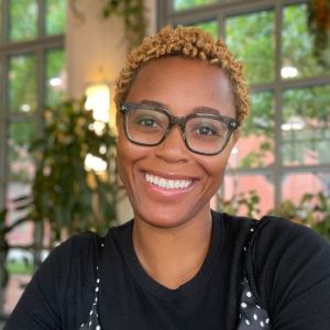 A headshot of Allanté Whitmore, PhD, a black woman with short light dyed hair and large rectangular glasses. She has a big smile on her face. Allanté is wearing a polka dot dress over a black t-shirt.