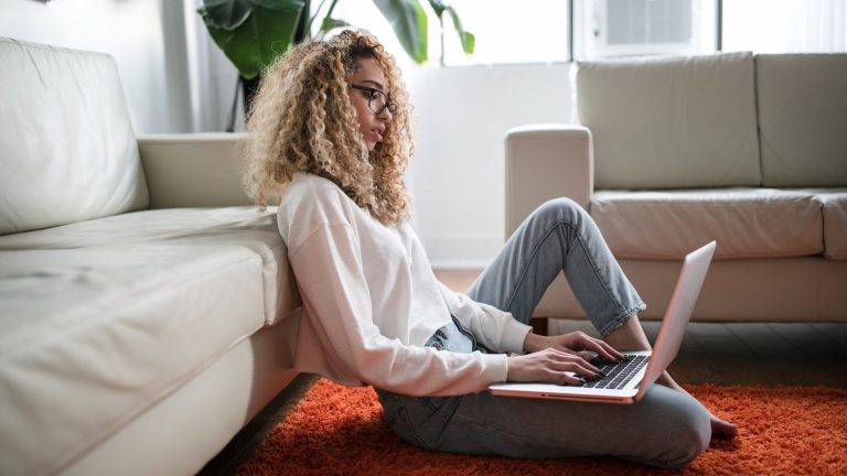 A black woman sits on the floor in front of a couch with her laptop resting on her knee while typing. The woman is wearing glasses, a white long sleeved shirt, and cropped jeans. The carpet in the living room is shaggy, orange, and looks comfy to sit on.