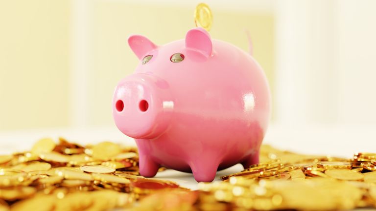 A pink piggy bank sits on a pile of gold coins. A gold coin is being dropped into the piggy bank by someone out of frame.