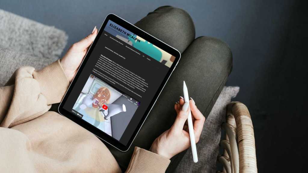 A woman sits on a chair holding a tablet. She has an e-pen in one hand to use to navigate on the screen. The tablet has Dr. Elizabeth McAplin's website pulled up on her Educational Design Samples page on her personal academic website.