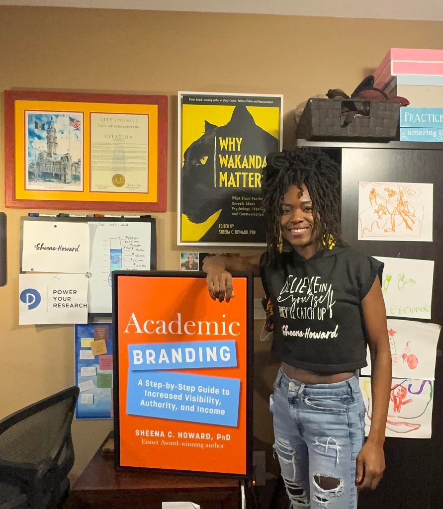 Professor Sheena C. Howard stands by a poster of her book, Academic Branding: A Step-by-Step Guide to Increased Visibility, Authority, and Income in her office. On the wall is a poster for her award-winning book Why Wakanda Matters. There's also a logo for her company, Power Your Research.
