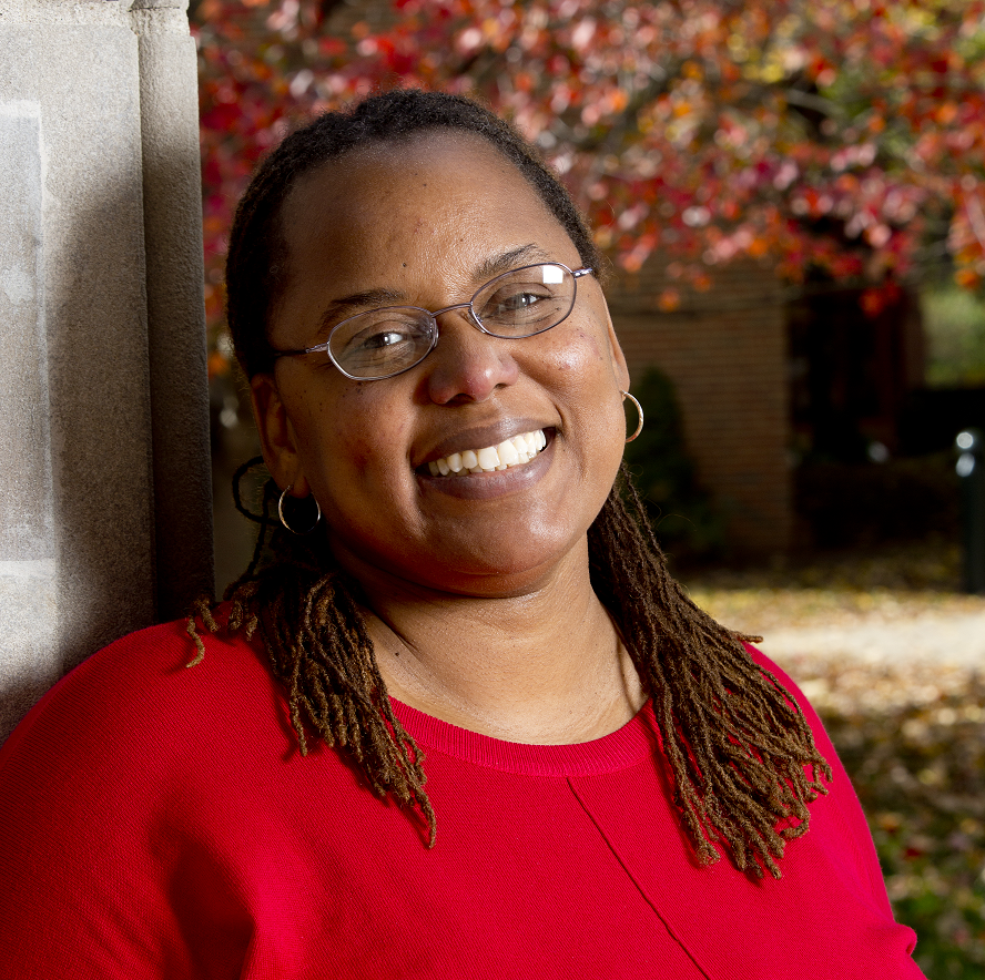 Carlotta Berry wears a red sweater, small hoop earrings, and wire frame glasses. She's smiling while leaning lightly against a column at the university. Behind her are the red leafed branches of a Japanese maple tree out of focus.