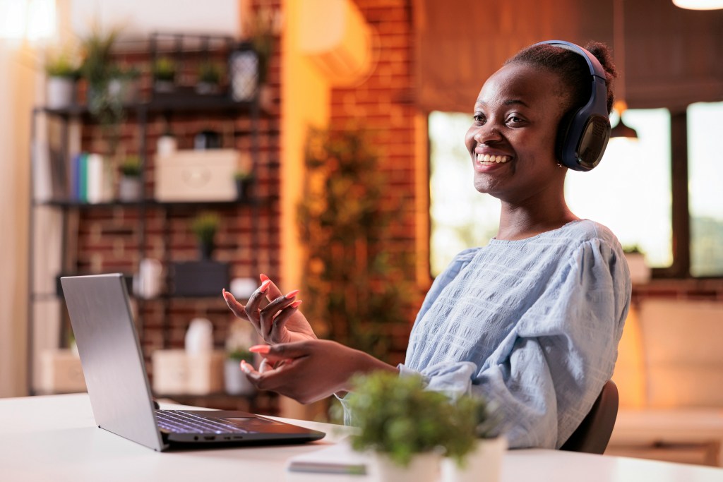 A black woman with headphones and a light blue dress sits at a desk in front of her open laptop. She's talking and smiling while presenting on camera.