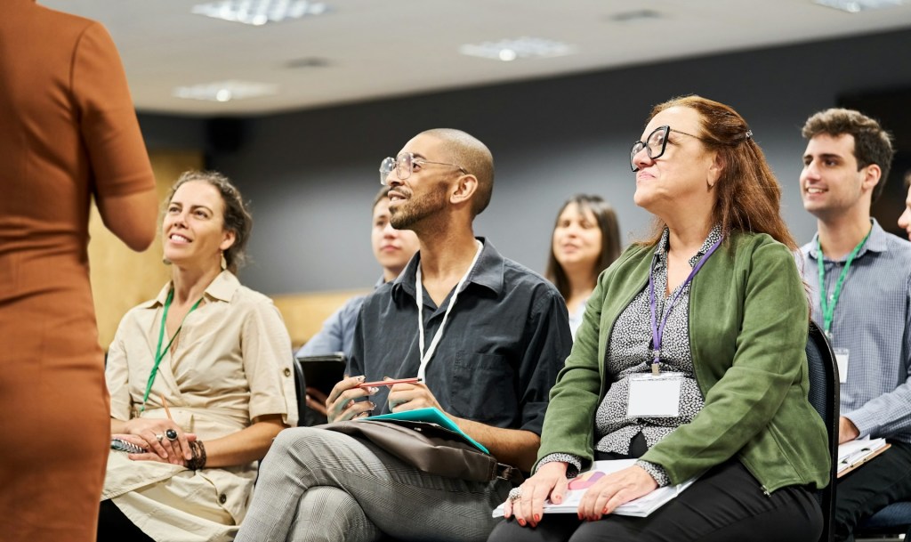 Six people smile at a speaker at a conference or talk. They're sitting in 3 rows in a smaller room or meeting space.
