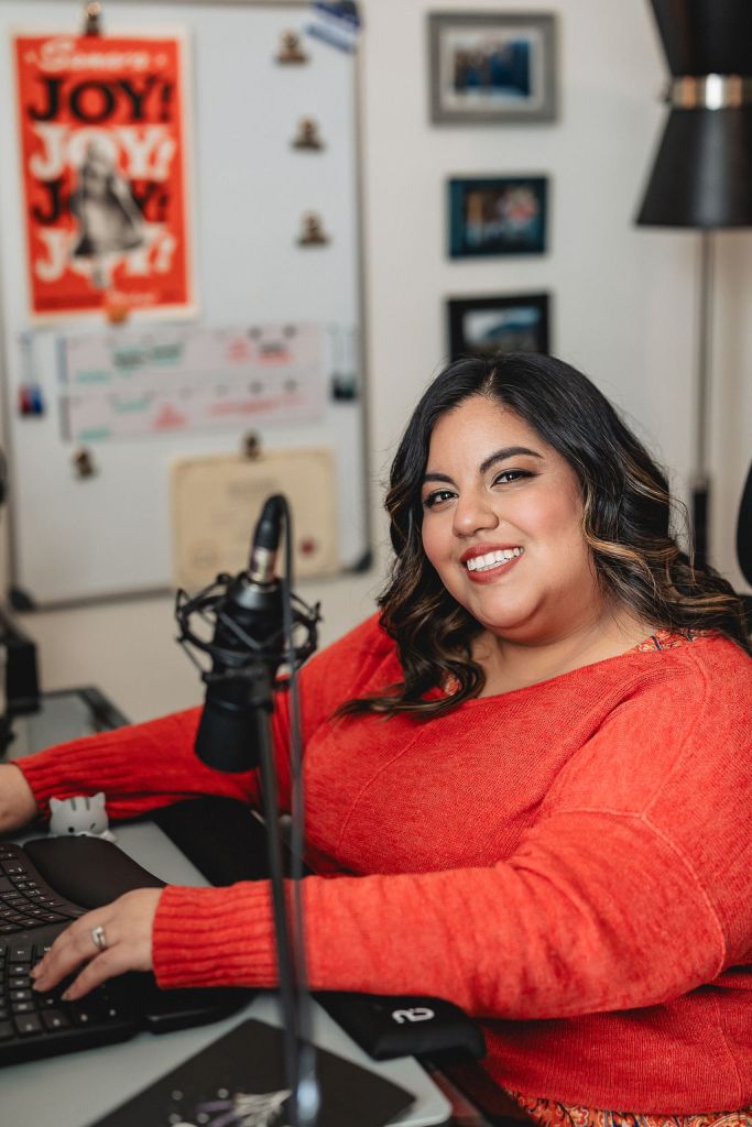 Jennifer van Alstyne sits at her desk in her home office in San Diego. She's a plus-sized latina woman with a pretty smile and long black hair. A professional microphone for podcast recording leans into the picture.