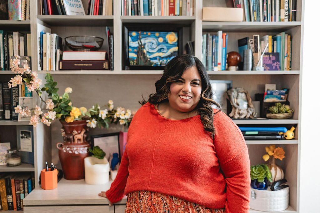 Jennifer van Alstyne stands in front of a built-in bookcase in her home office. There is a llama vase with flowers, and a Van Gogh LEGO set behind her on the shelves filled with books.