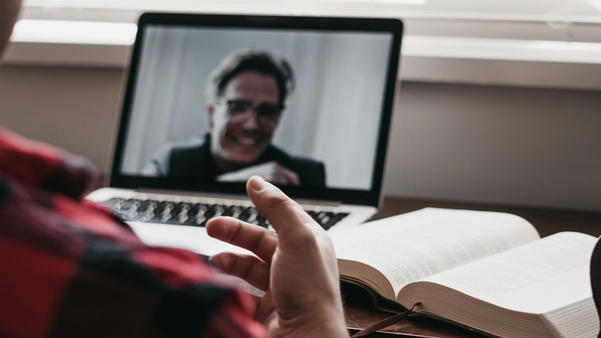View over the shoulder of a professor who is attending a live virtual event. On his open laptop screen is the speaker, a person with short hair and glasses, smiling.