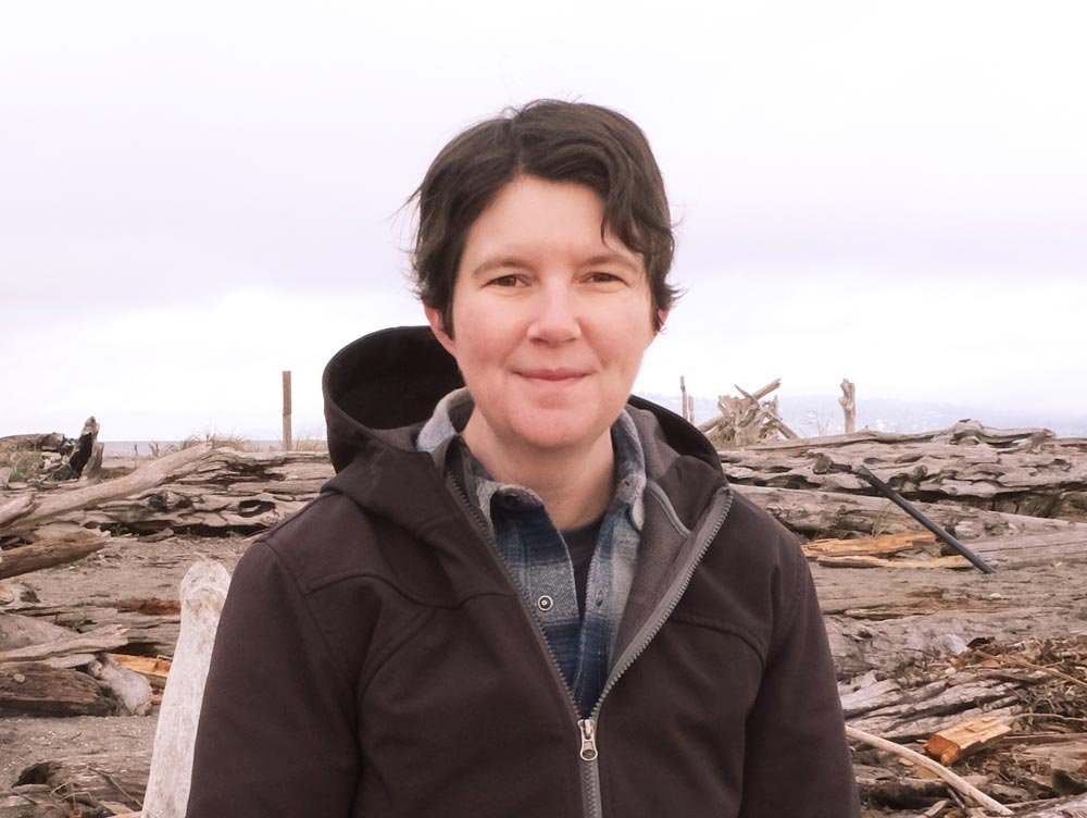 HR Hegnauer on a driftwood beach on a cloudy day. She's wearing a brown zip up jacket and plaid shirt, smiling.