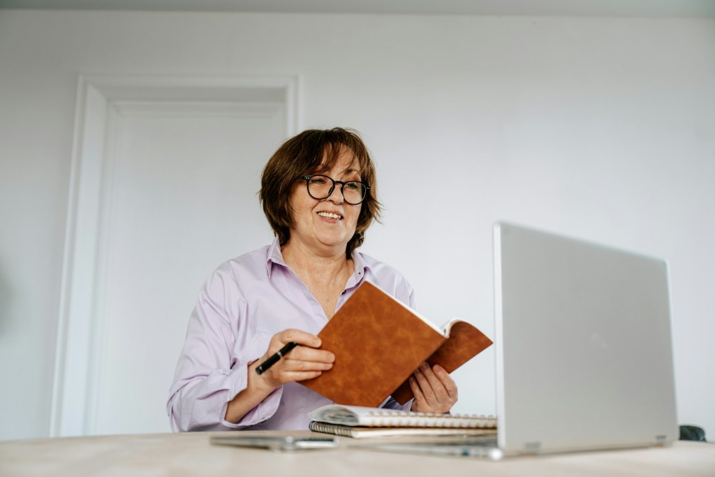 A middle-aged woman wearing a lilac button down top holds a notebook and pen. She's smiling at the open laptop on her desk as if she's on a video call.