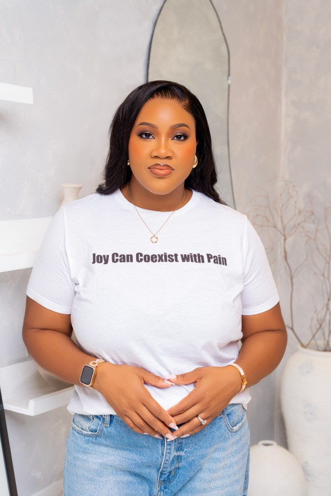 Chinasa Elue wears a white t-shirt and jeans. The shirt has black text that reads, "Joy Can Coexist with Pain".