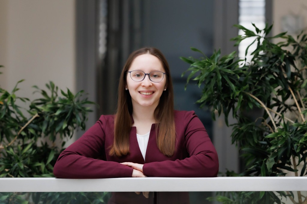 Ana Rebeka Kamšek wears a maroon blazer and glasses. She's smiling warmly while leaning forward against a balcony railing.