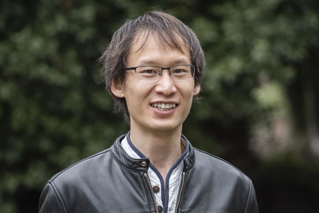 A man with short brown hair and glasses smiles outdoors. He is wearing a black leather jacket over a white shirt. The background is blurred greenery, suggesting a park or garden setting.