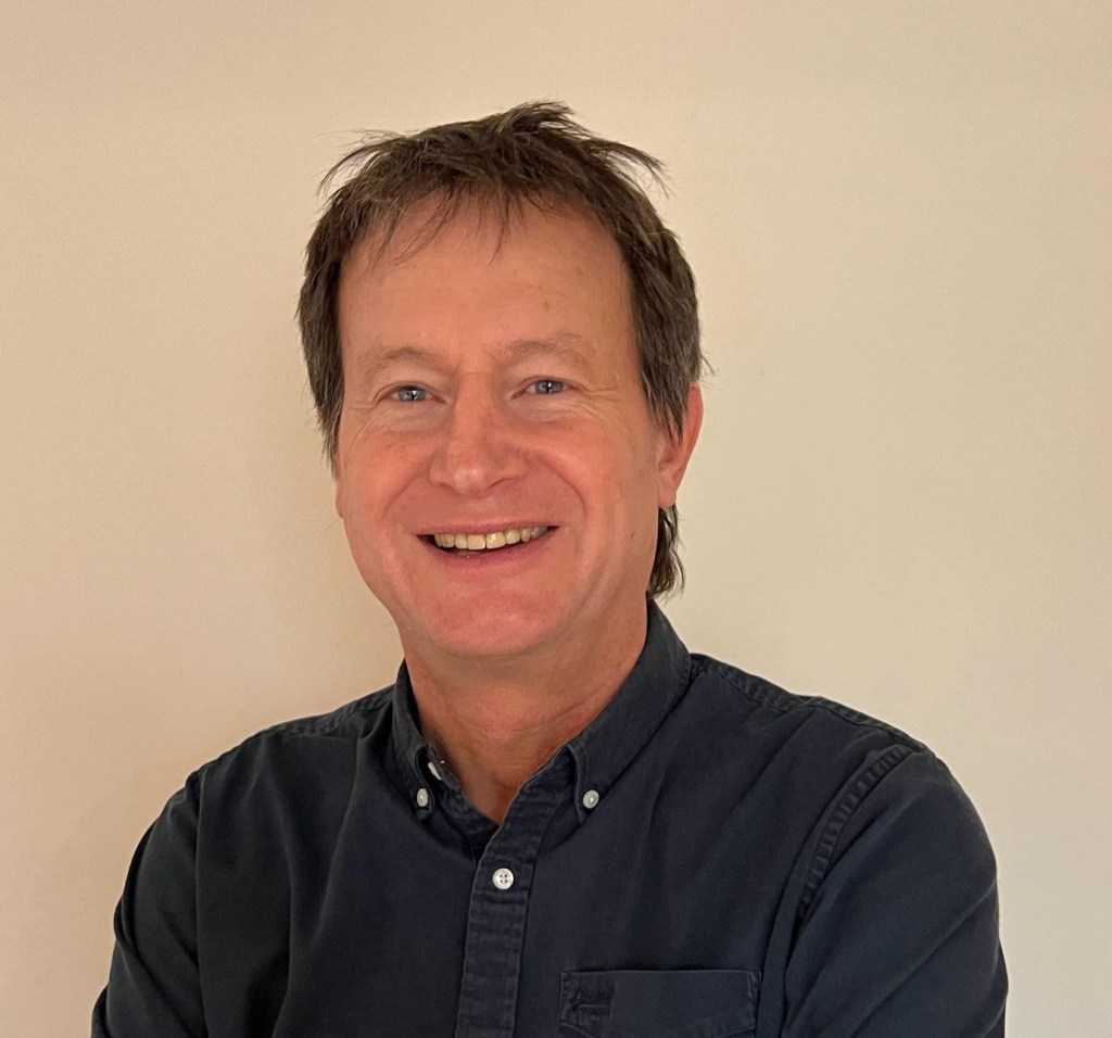 Professor Bruce Hood, a man with short, brown hair smiles at the camera against a plain, off-white wall. He is wearing a dark blue, button-down shirt.